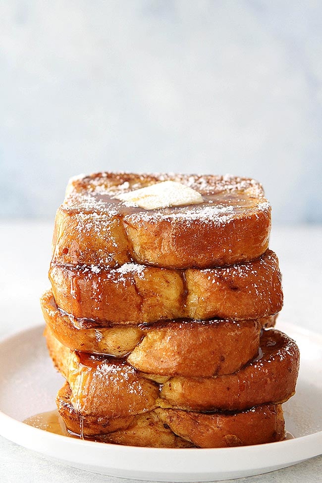 Stack of brioche French toast on plate with butter, syrup, and powdered sugar. 