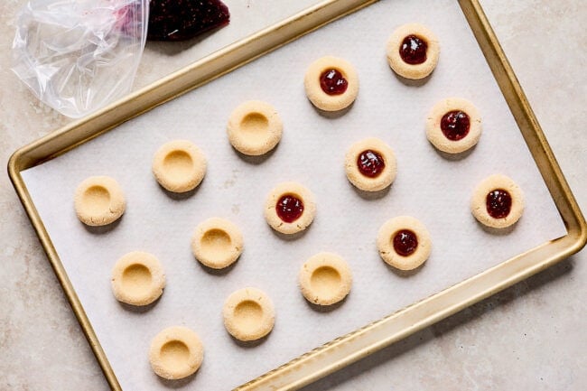 thumbprint cookie dough balls on baking sheet being filled with raspberry jam.